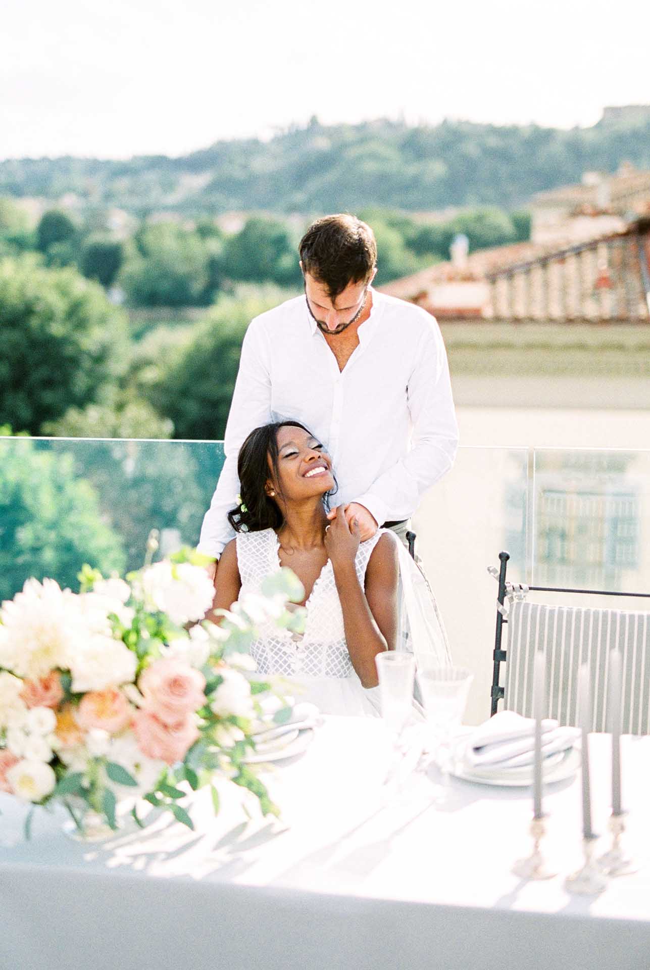groom hugs from behind bride sitting at the table on the terrace of the building
